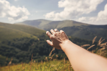 Hand with engagement ring reaching out to green hills in summer mountains. Summer vacation in mountains. Silver ring with crystal. View on top of green hills and clouds