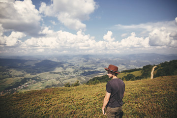 Naklejka na ściany i meble Stylish hipster traveler in hat standing on top of hills on background of summer mountains. Copy space. Travel and wanderlust concept.