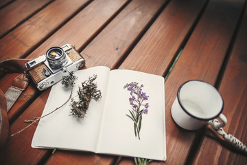 Travel and wanderlust image. Photo camera, mug, notebook,gathered herbs and wildflowers on wooden rustic background. Summer travel essentials in mountains