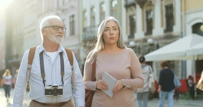 Senior two male and female tourists with photocamera and map stepping the city square and talking. Outside.