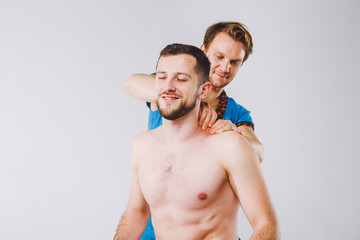 Theme massage and body care. A handsome Caucasian male doctor in blue uniform and beard diagnosing muscles of neck and shoulders, stretching guy with good figure on massage table a white background