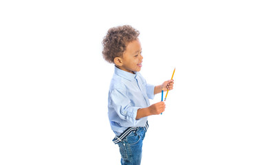 A portrait of a little dark-skinned boy with curly hair in jeans and a light-colored shirt is standing in profile and holding pencils playfully