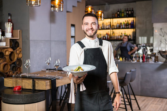 A handsome young guy with a beard dressed in an apron standing in a restaurant and holding a white plate with a moth. Against the background, the bar counter and the loft style interior