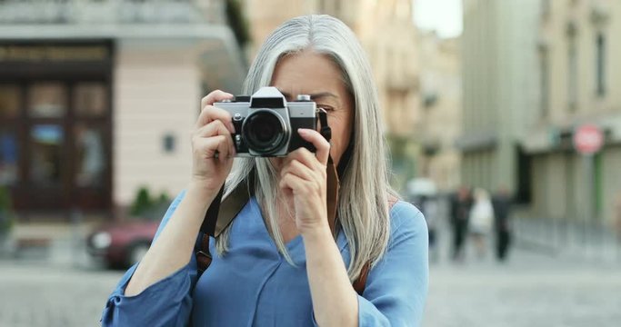 Portrait of the gray haired Caucasian senior woman with long hair standing in front of the camera at the street and taking photo on the photocamera. Close up. Outdoor.