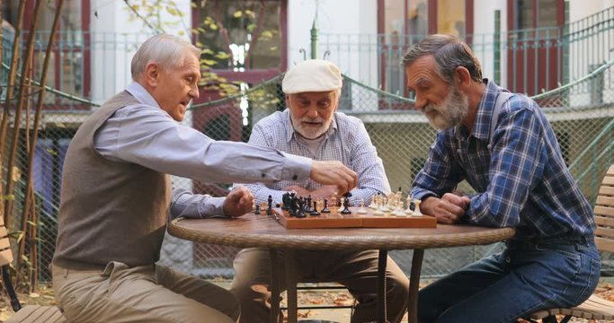 Caucasian old grey haired men sitting at the chess game on the table and doing their chessmen moves. Outdoors.