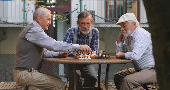 Portrait shot of the three senior Caucasian men on retirement sitting in the yard round the table and playing a chess game while moving figures. Outdoor.