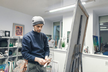 Naklejka na ściany i meble Portrait of a young artist is in a studio near an easel with a canvas with instruments in his hands. Artist with brushes in his hands draws a picture.