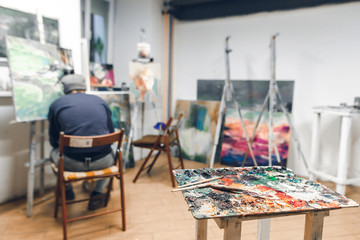 Palette and brush stand on the background of a painter sitting on a chair and paints a picture at...