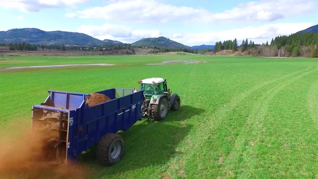 green farm tractor fertilizing large pece of farm land in beautiful BC surrounded by mountains and trees - shot from aerial drone in 60 frame per second