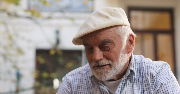 Close up of the senior good looking Caucasian man in a cap smiling and thinking while playing a chess game at the fresh air. Portrait. Outside.