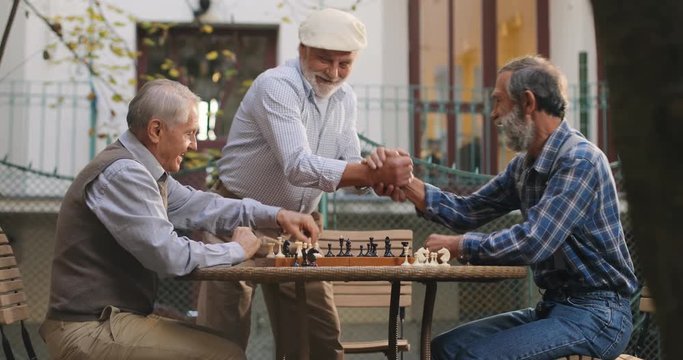 Caucasian cheerful senior man in a cap coming into the yard and joining his two friends neighbors in the chess game. Outside.Quarantine Concept Coronavirus Pandemic