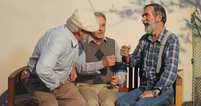 Two Caucasian Old Men On Retirement Sitting On The Bench And Chatting, Then The Third One Joining Them. Neighbors On A Sunny Day. Outside.