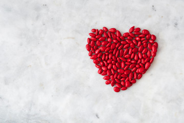 Fototapeta premium Red candy coated sunflower seeds in a heart shape on a white granite background for Valentine’s Day