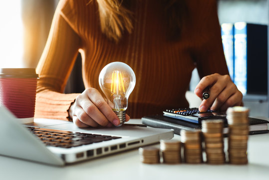 business man hand holding lightbulb with using smartphone and calculator to calculate and money stack. idea saving energy and accounting finance in morning light