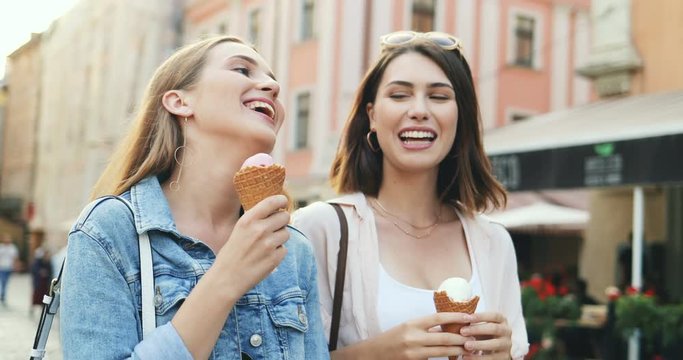 Two happy smiled young Caucasian women best friends walking the city street with ice-cream in hands and having fun. Outside. Close up.