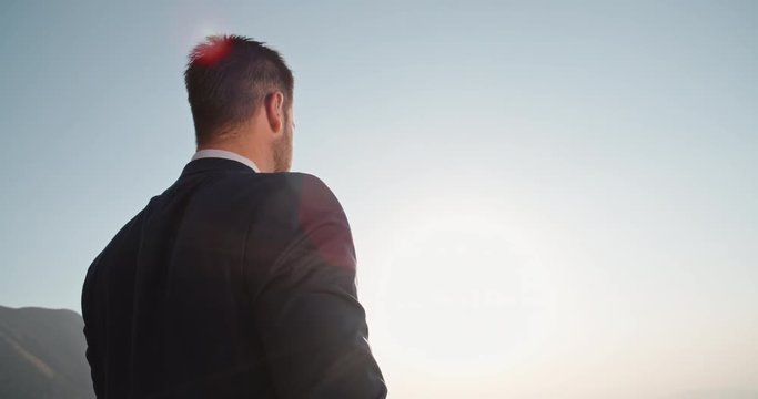 Young confident entrepreneur in black suit standing on top of a mountain, looking far at sunset - way to success, on top of the world concept 4k