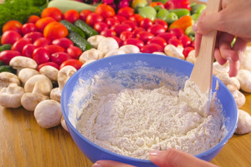 Woman's hands is kneading the yeast dough with a wooden spatula in blue bowl on the table with vegetables.
