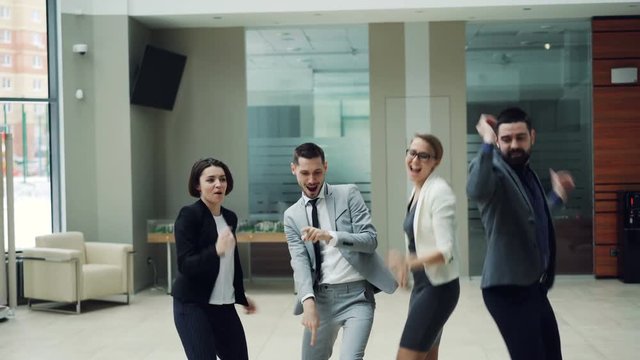 Group of excited young people is dancing in hall of modern business center enjoying corporate party and having fun. Men and women are wearing smart suits.