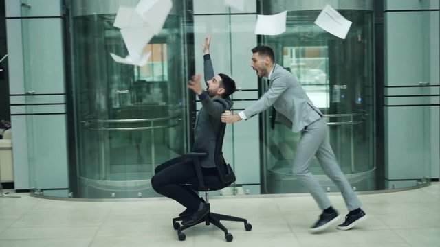 Slow motion of happy business partners having fun in office riding chair and throwing papers contracts enjoying party. Enjoyable job and youth concept.