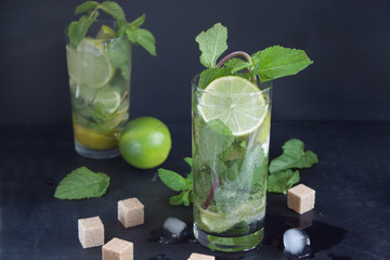 Two glasses of mojito cocktail with fresh lime and mint on a dark background. Ice cubes and pieces of brown sugar are lying on a table.