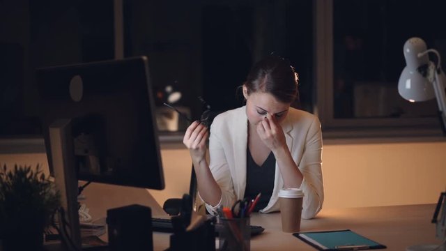 Pretty young woman secretary is working on computer late at night then taking off glasses and rubbing tired eyes and face then drinking to-go coffee.