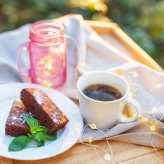Close-up of cup of tea with a brownie cake decorated with some mint on a wooden table outdoors. Mason jar decorated with lights on the background
