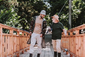 Pleasant aged fisherman walking along the bridge with his son while having a lively talk