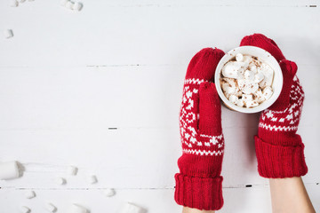 Female hands holding hot chocolate with marshmallow above white wooden table. Top view