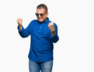 Middle age arab man wearing sunglasses over isolated background very happy and excited doing winner gesture with arms raised, smiling and screaming for success. Celebration concept.