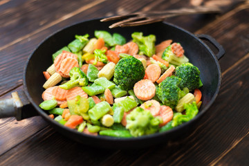 A mixture of frozen different vegetables in a pan on a wooden background. - obrazy, fototapety, plakaty