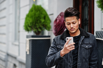 Handsome and fashionable indian man in black jeans jacket posed outdoor and looking at mobile phone.