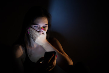 Teen girl excessively sitting at the phone at home. he is a victim of online social networks. Sad teen checking phone sitting on the floor in the living room at home with a dark background.