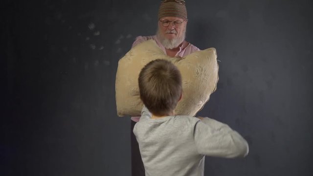 Concept of gymnastics and sport at home. Elderly retired father with gray beard and in pink T-shirt holds pillow and trains boy for boxing. Brave child beats on pillow with all his might.