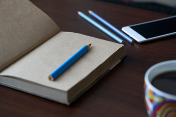 Recycled paper notebook, smartphone, blue pencil and a cup of coffe on a dark table. Work desk with notepad, mobile phone and a pencil.