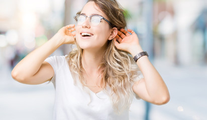 Beautiful young blonde woman wearing glasses over isolated background Smiling pulling ears with fingers, funny gesture. Audition problem