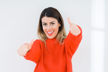 Young woman wearing casual red sweater over isolated background approving doing positive gesture with hand, thumbs up smiling and happy for success. Looking at the camera, winner gesture.