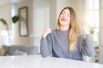 Young beautiful woman wearing winter sweater at home very happy and excited doing winner gesture with arms raised, smiling and screaming for success. Celebration concept.