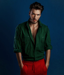 Fashion portrait of young man in green shirt and red pants poses over dark blue wall with contrast...