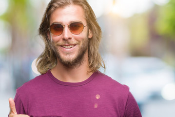 Young handsome man with long hair wearing sunglasses over isolated background doing happy thumbs up gesture with hand. Approving expression looking at the camera with showing success.