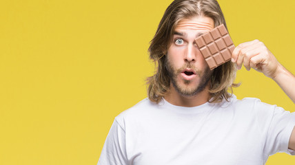 Young handsome man with long hair eating chocolate bar over isolated background scared in shock...