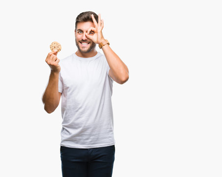 Young handsome man eating chocolate chips cookie over isolated background with happy face smiling doing ok sign with hand on eye looking through fingers