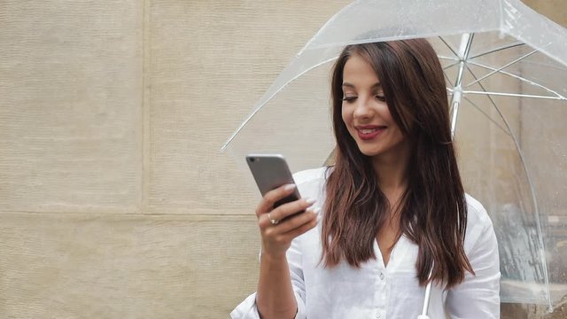 Beautiful young business woman using smartphone on the street in rainy weather, smiling, holding umbrella, communication concept
