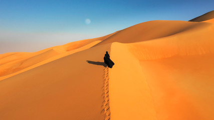 Aerial view from a drone flying next to a woman in abaya (United Arab Emirates traditional dress) walking on the dunes in the desert of the Empty Quarter. Abu Dhabi, UAE. © Kertu