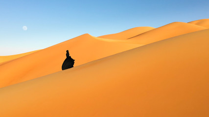 Fototapeta premium Aerial view from a drone flying next to a woman in abaya (United Arab Emirates traditional dress) walking on the dunes in the desert of the Empty Quarter. Abu Dhabi, UAE.