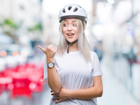 Young blonde woman wearing cyclist security helmet over isolated background smiling with happy face looking and pointing to the side with thumb up.