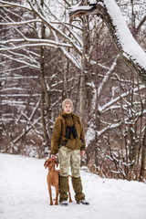 Female hunter in camouflage, armed with a rifle, standing in a snowy winter forest with duck prey