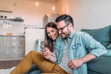 Couple watching at smart phone together while sitting on the floor in a living room at home.