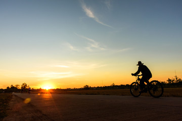 Silhouette of an old man riding a bicycle at sunset in public park.Orange blue sky on background.