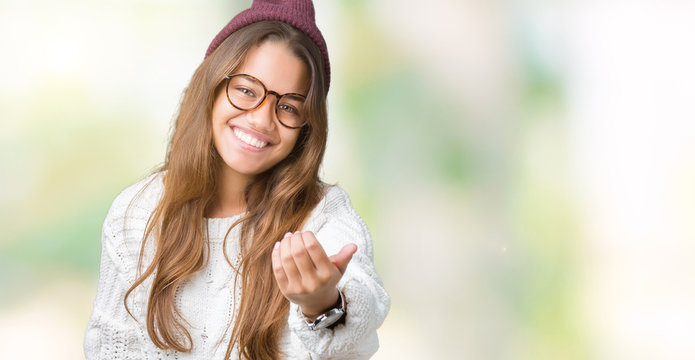 Young beautiful brunette hipster woman wearing glasses and winter hat over isolated background Beckoning come here gesture with hand inviting happy and smiling