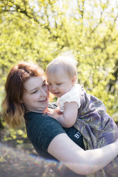Mother smiling while wrapping her baby daughter in a sling for babywearing. Lifestyle image shot on location in a park on a sunny spring day. 